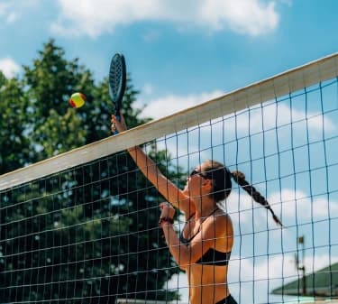 A foto foi feita em uma quadra de beach tennis. Nela, há uma mulher praticando a modalidade, enquanto salta acima da altura da rede e rebate a bolinha com uma raquete. #ParaTodosVerem