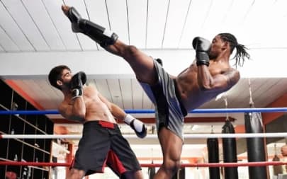 Na foto, dois homens estão em ringue de lutas, sendo diferenciados pela cor do shorts de cada um. Os dois estão praticando kickboxing, enquanto um ataca e o outro se defende. #ParaTodosVerem