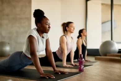 A foto mostra três mulheres em um estúdio de yoga. Elas estão em uma aula, deitadas cada uma em seu próprio tapete, fazendo o alongamento. Há também uma bola ao fundo que é um acessório utilizado durante a prática. #ParaTodosVerem